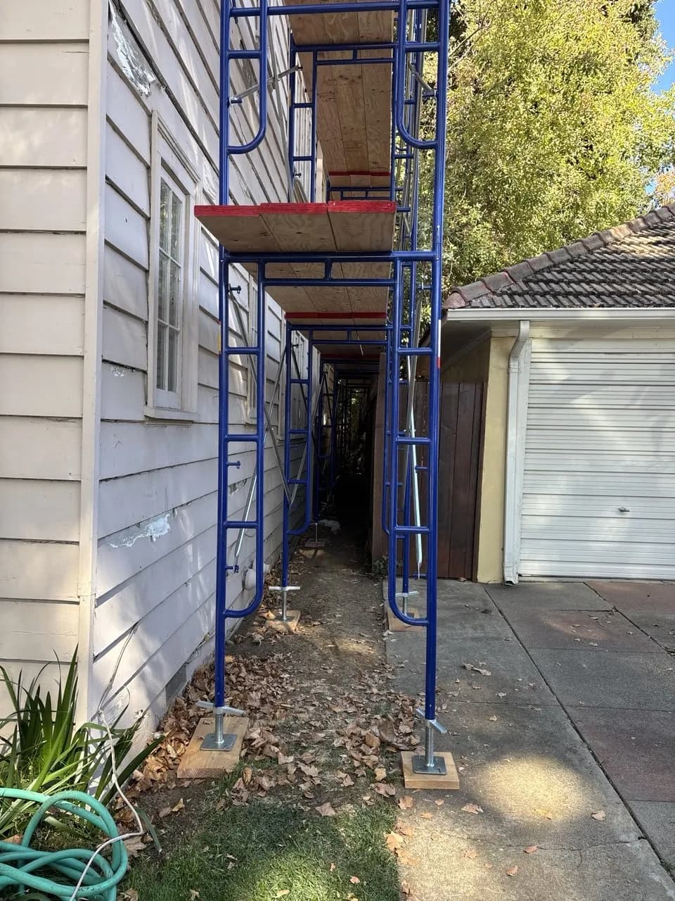 Blue frame scaffold on screw jacks and wood mudsills along a side yard, with planks on two levels